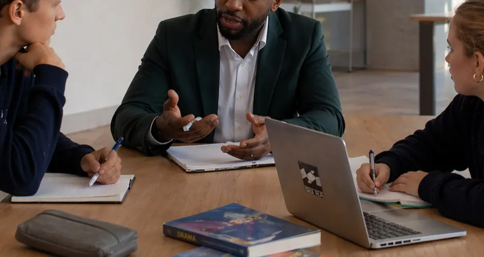 A tutor talking with two students around a table with notebooks and a laptop